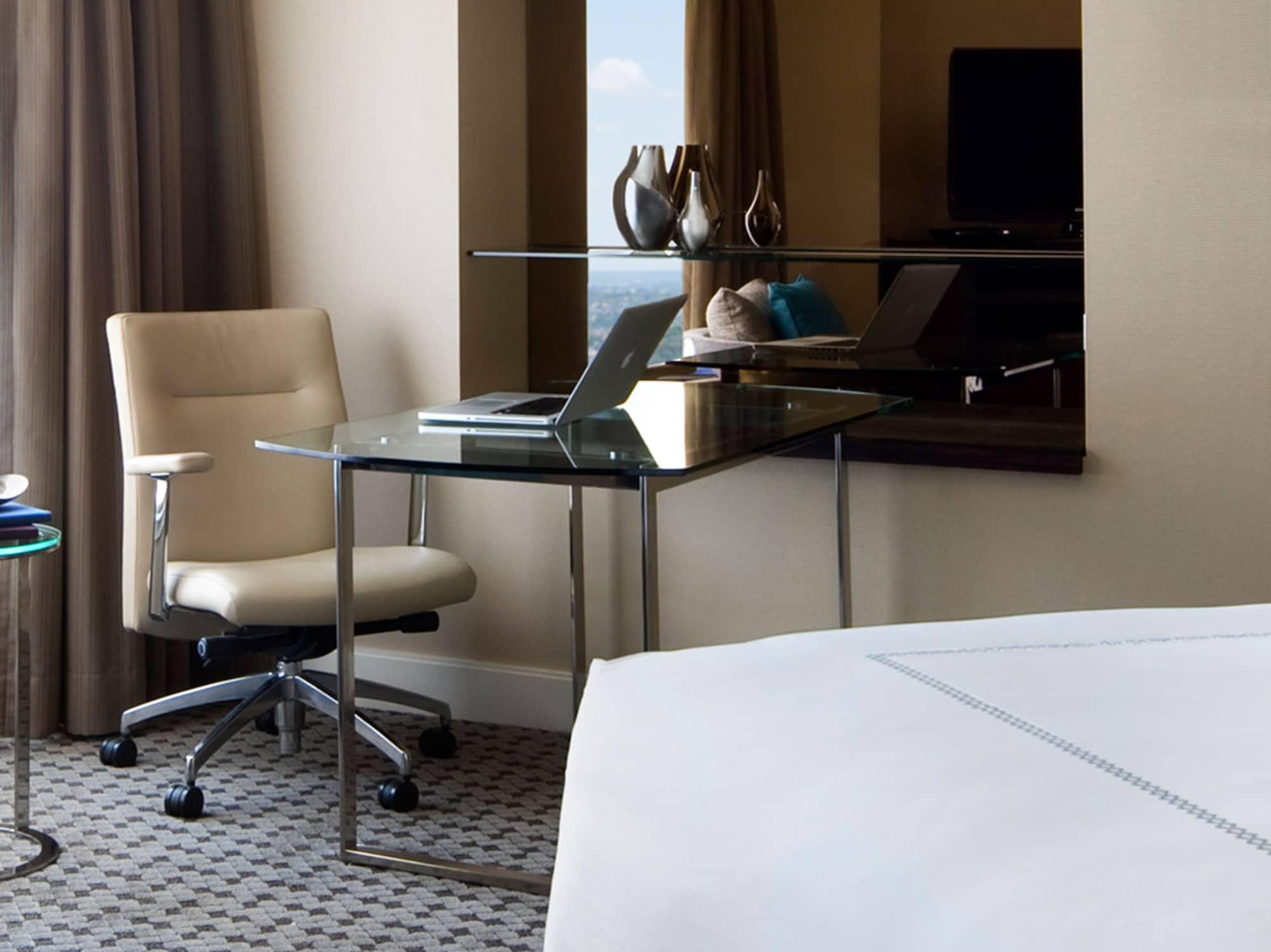Four Seasons Hotel Sydney Kültér fotó The photo depicts a modern hotel room setup. On one side, there's a sleek glass desk with a laptop set up on it, accompanied by a comfortable, ergonomic chair. The desk is positioned next to a window draped with curtains, and there are decorative ite