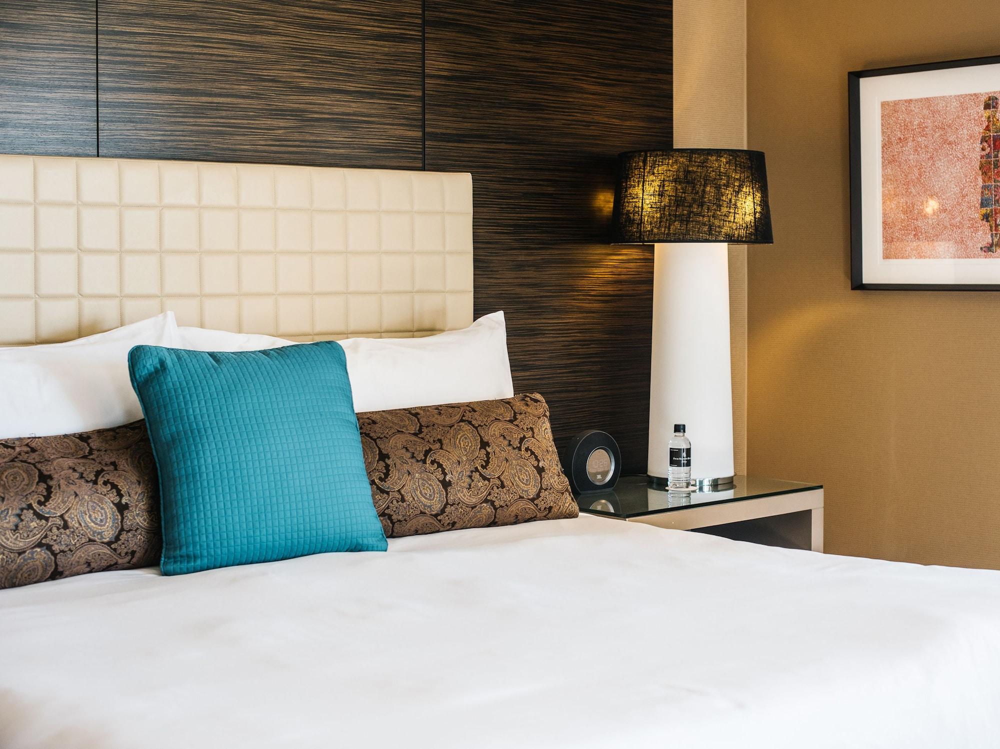 Four Seasons Hotel Sydney Kültér fotó The photo shows a neatly arranged bed in a hotel room. The bed features a white duvet and a couple of decorative pillows, including a brown patterned one and a solid turquoise one. Behind the bed, there is a modern headboard with a grid pattern. Next