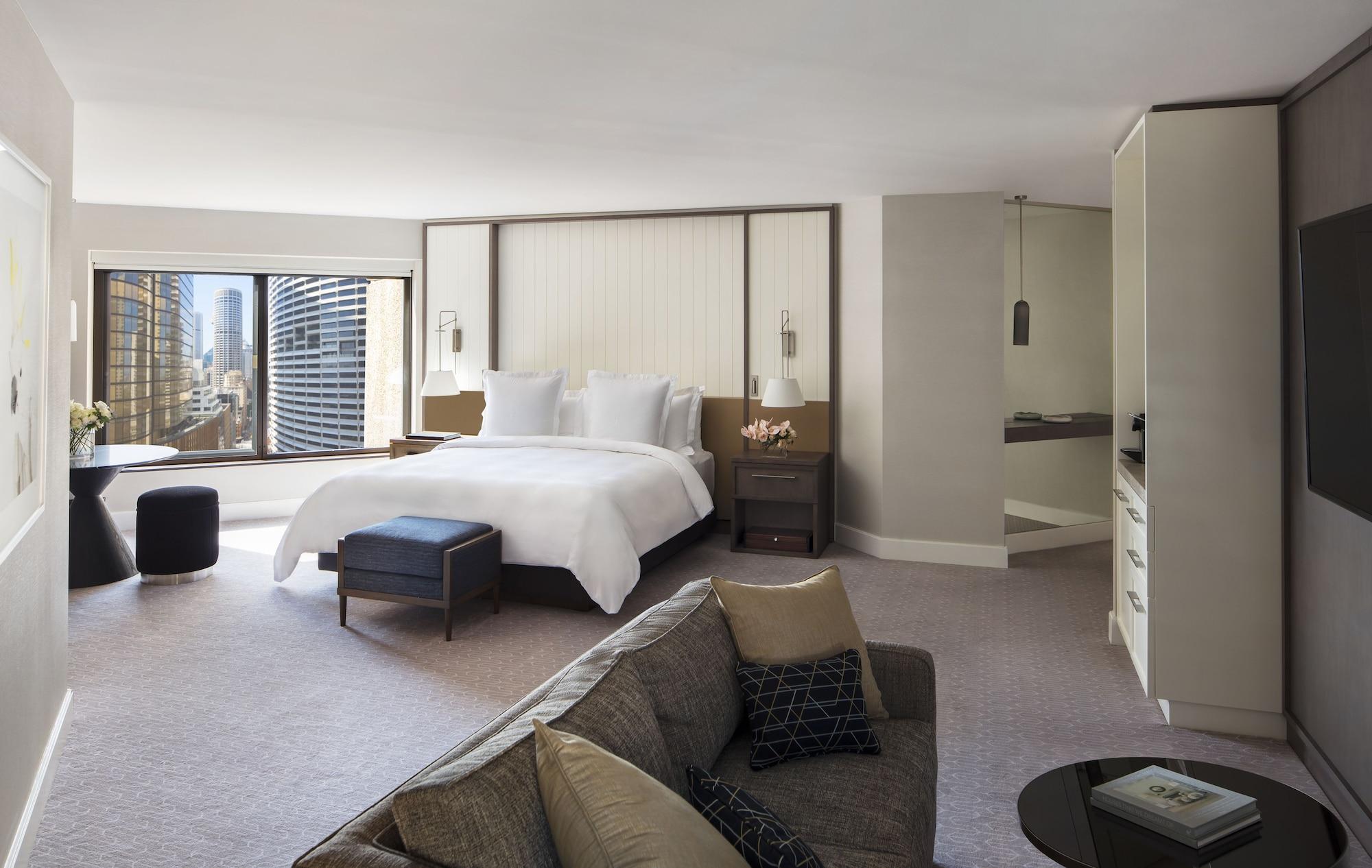 Four Seasons Hotel Sydney Kültér fotó The photo shows a modern hotel room featuring a spacious layout. In the center, there's a large bed with white linens, flanked by nightstands with lamps. To one side, a comfortable sofa is positioned, complemented by a small coffee table. There is a 