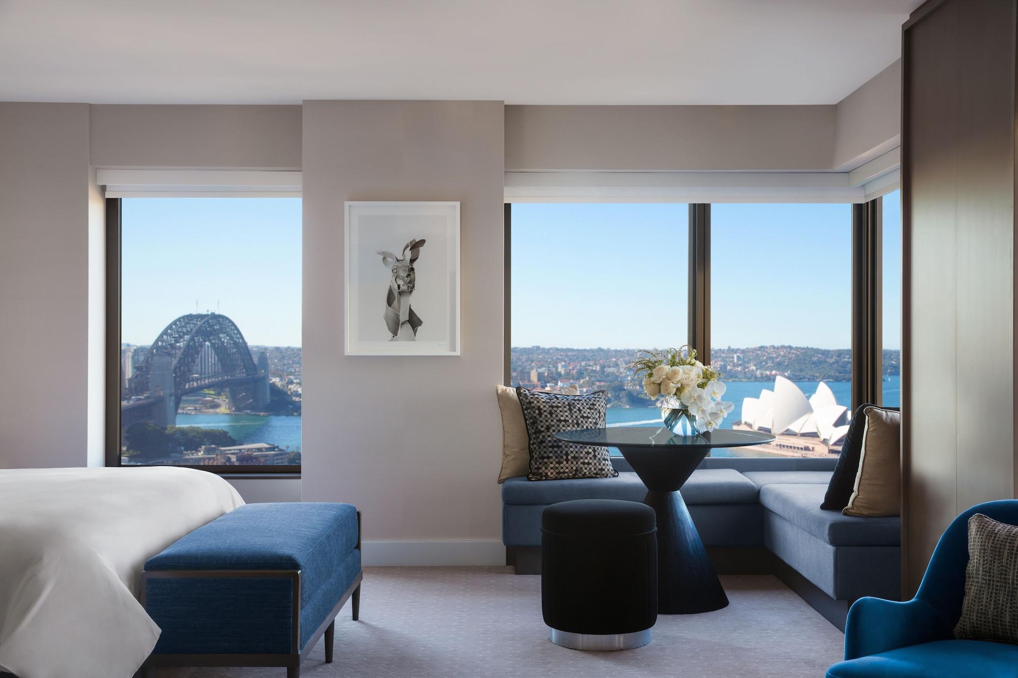 Four Seasons Hotel Sydney Kültér fotó The photo shows a modern hotel room with large windows offering a view of the Sydney Opera House and the Sydney Harbour Bridge. The interior features a comfortable bed with a blue upholstered bench and decorative pillows. There's a round black table 