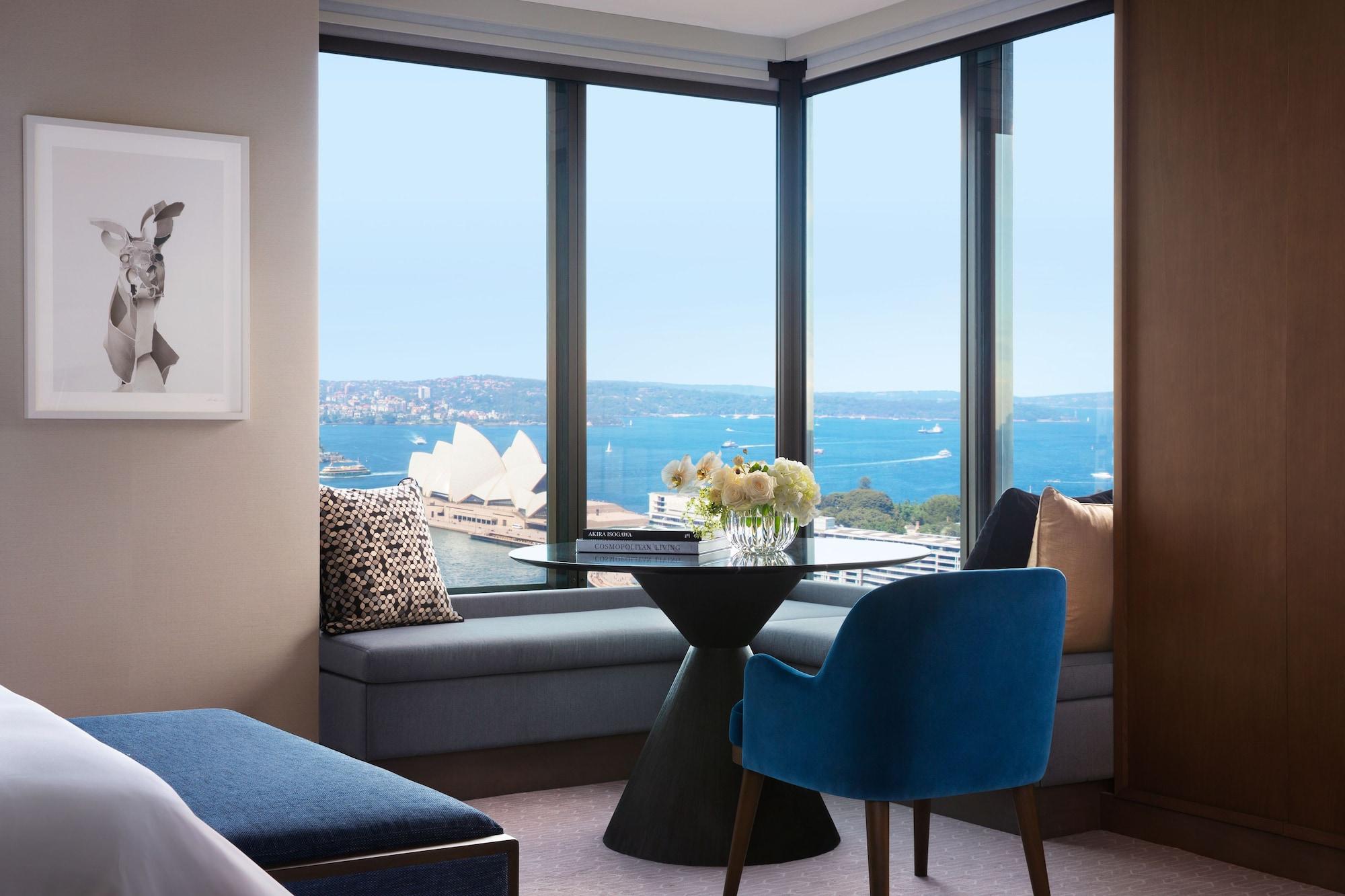 Four Seasons Hotel Sydney Kültér fotó The photo shows a stylish hotel room with a large window offering a view of the water and the Sydney Opera House in the distance. There is a small, modern dining table with a vase of flowers on it, situated near the window. A comfortable blue chair i