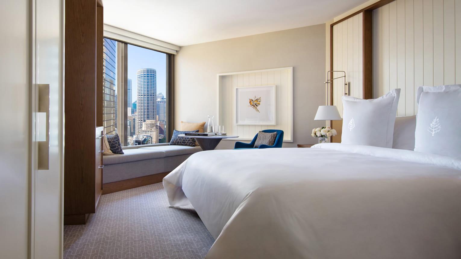 Four Seasons Hotel Sydney Kültér fotó The photo shows a modern hotel room featuring a comfortable bed with crisp white linens. There is a well-lit area with a large window that offers a view of the city skyline. A cozy seating nook can be seen near the window, with a small table and chai