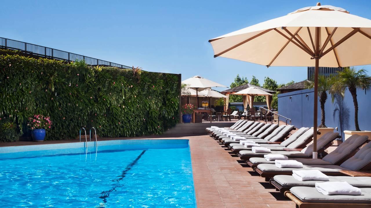 Four Seasons Hotel Sydney Kültér fotó The photo shows a beautiful outdoor pool area. The pool has clear blue water and is lined with comfortable lounge chairs. Some of the chairs are shaded by large umbrellas. In the background, there are more umbrellas and seating areas, suggesting a re