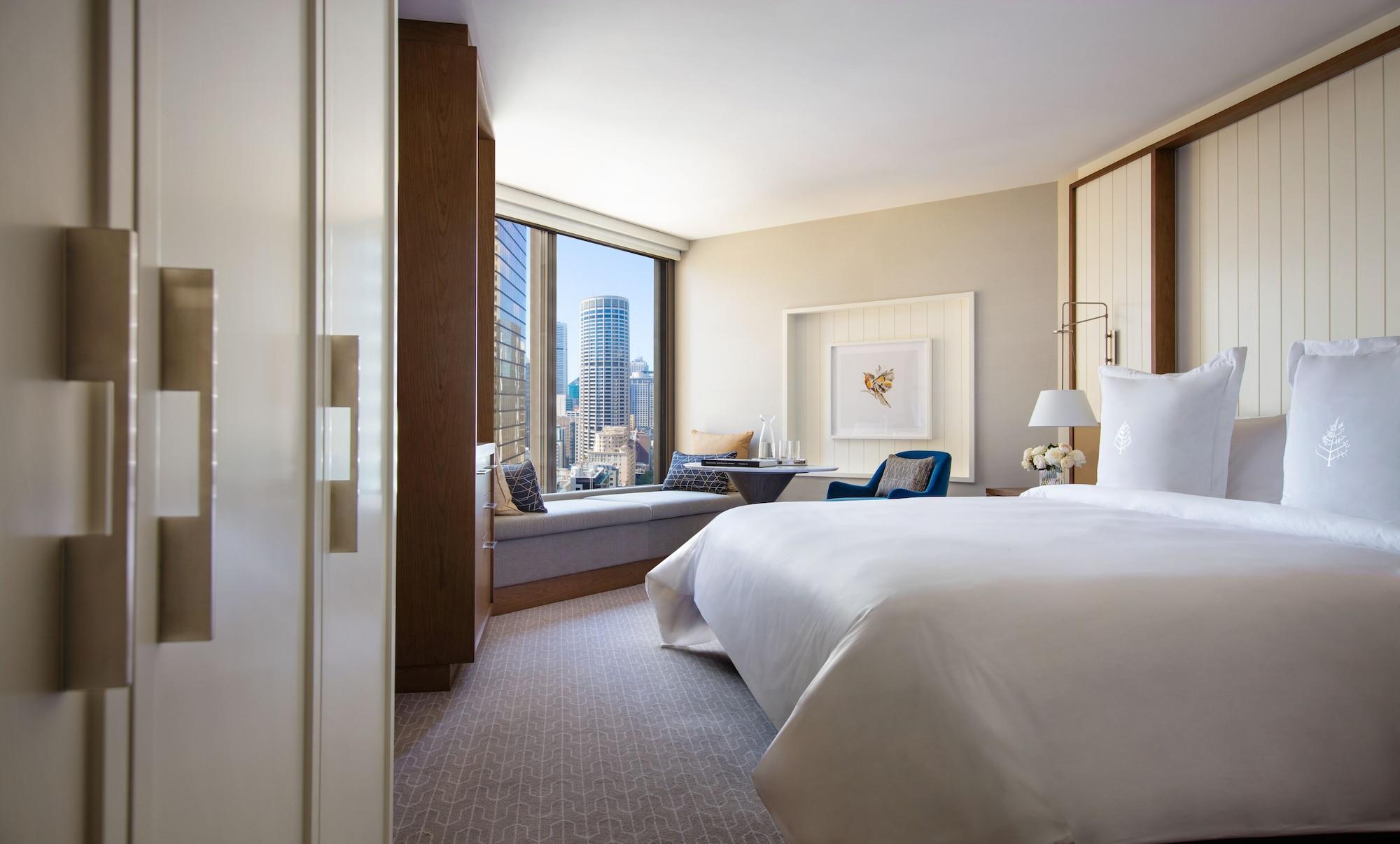 Four Seasons Hotel Sydney Kültér fotó The photo shows a modern hotel room with a large bed, featuring a crisp white duvet and pillows. There is a stylish seating area with a cozy window nook and comfortable chairs, allowing for a view of the city skyline outside. The room has contemporar