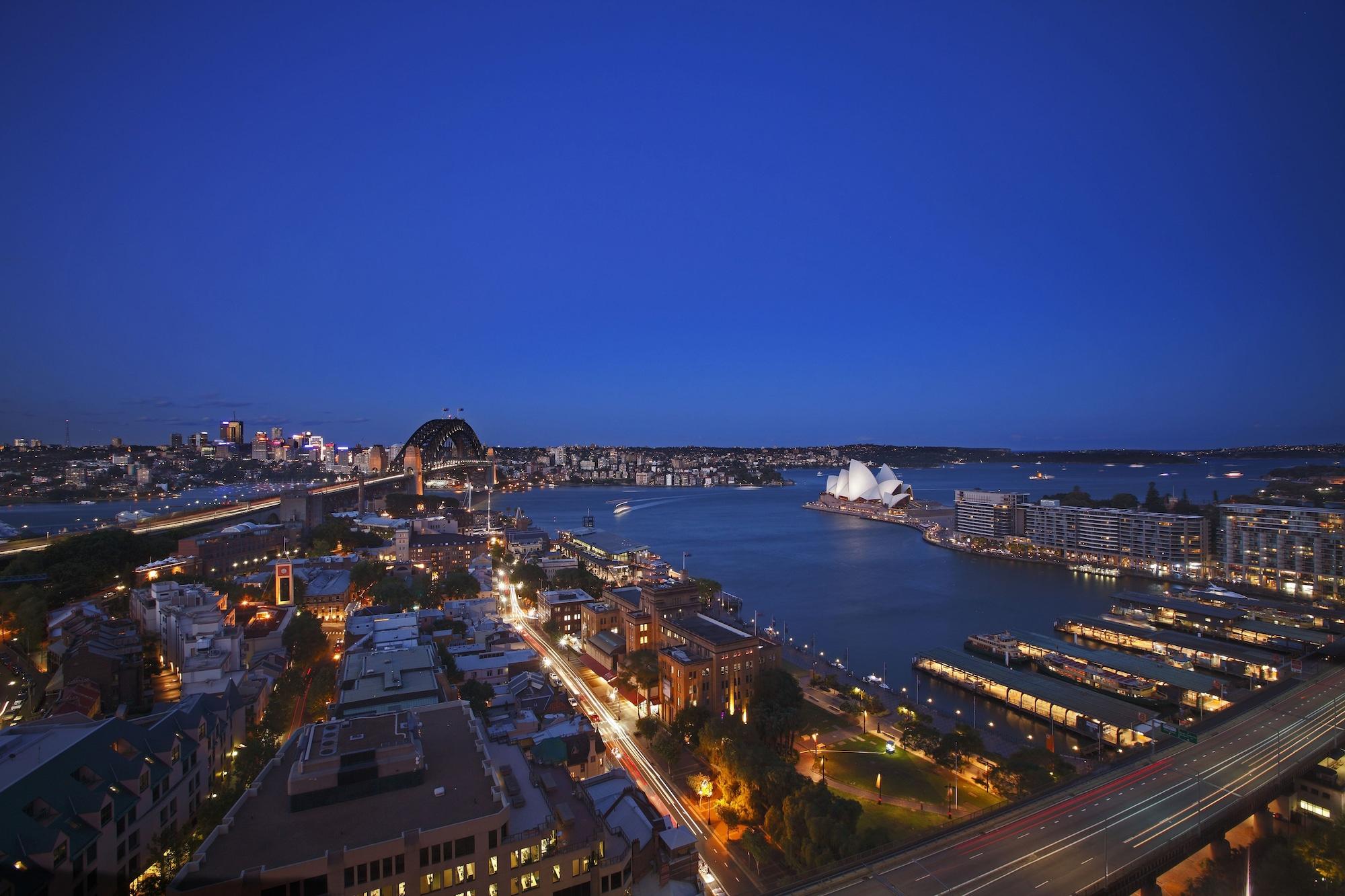 Four Seasons Hotel Sydney Kültér fotó The photo captures a scenic view of Sydney, Australia, during twilight. The iconic Sydney Opera House is prominently visible along the waterfront, characterized by its distinctive white sail-like roof. In the background, the silhouette of the Sydney 