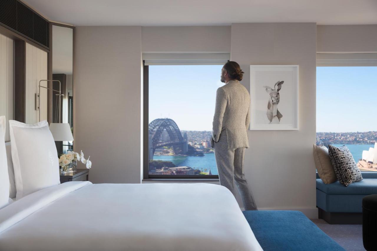 Four Seasons Hotel Sydney Kültér fotó The photo features a modern hotel room with a large bed and a stylish decor. A person dressed in a formal outfit stands near a window, looking out. The view includes a scenic landscape, possibly of Sydney, showcasing the harbor and a landmark structu